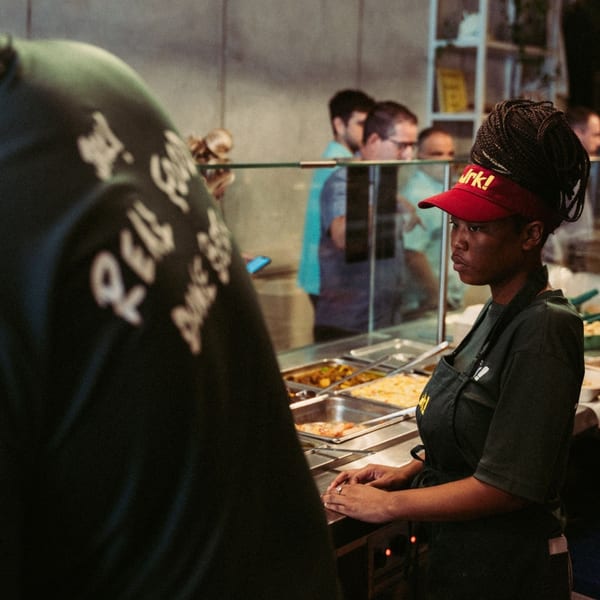 A Jrk! employee in a red visor and black apron stands behind a food counter with customers.