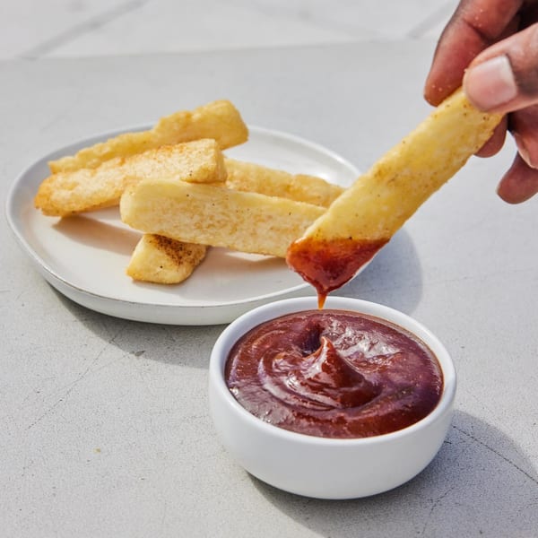 A hand dips a golden-brown Jrk! Jamaican fry into a small white bowl of rich, dark red sauce.