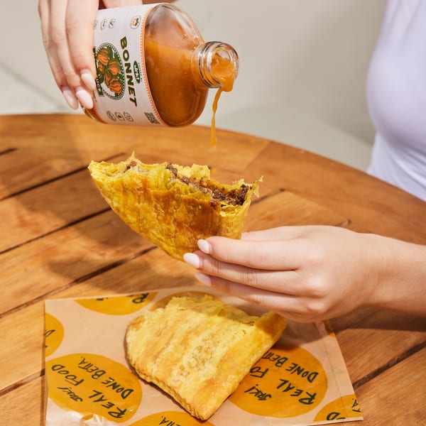 A person pours Jrk! Bonnet sauce onto a Jamaican beef patty, with another patty on branded paper.