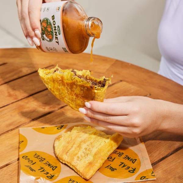 Hands pouring Jrk! Bonnet hot sauce onto a Jamaican beef patty with a bite taken out.