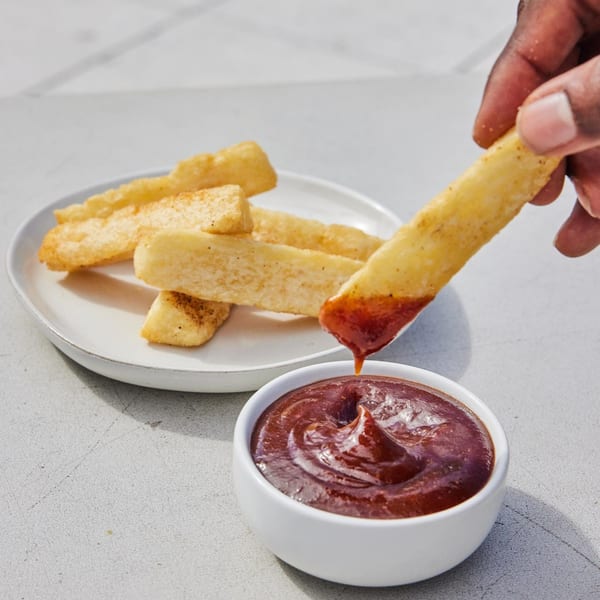 A hand dips a golden-brown fry into a small white bowl of dark red jerk BBQ sauce.