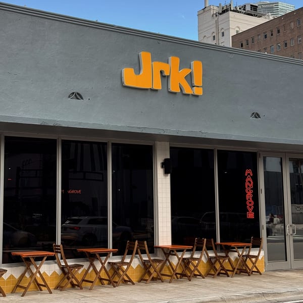 Exterior of Jrk! restaurant with orange sign, large windows, and outdoor seating with wooden tables and chairs.