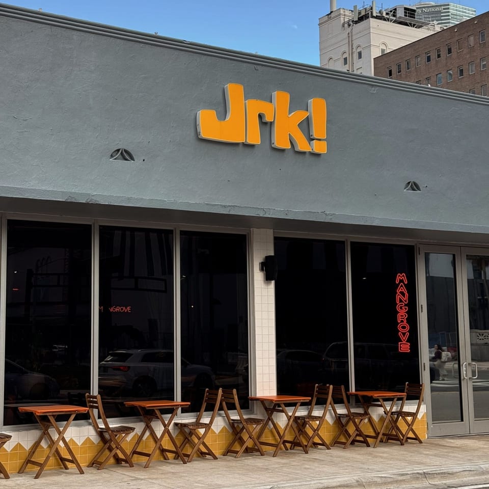 Exterior of Jrk! restaurant with orange sign, large windows, and outdoor seating with wooden tables and chairs.
