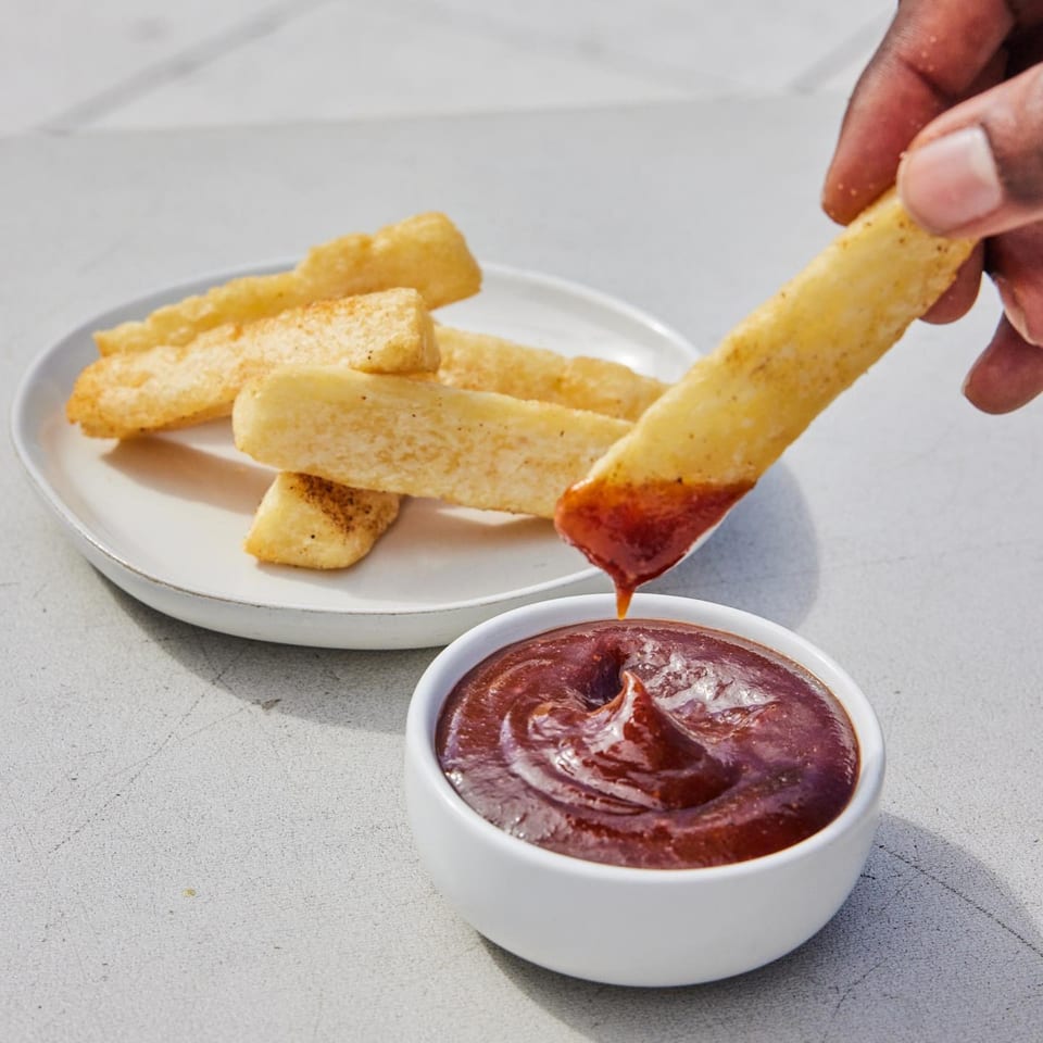 A hand dips a golden-brown Jamaican festival dumpling into a small white bowl of BBQ sauce.