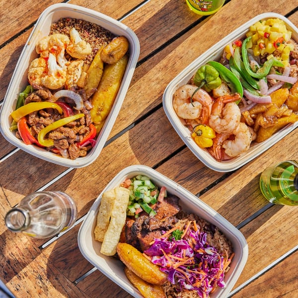 Three Jrk! Jamaican takeout bowls with various meats, vegetables, rice, and plantains on a wooden table.