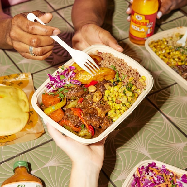 Hands holding a Jrk! Jamaican oxtail meal with rice, plantains, corn, and slaw.