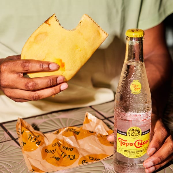 A person holds a partially eaten Jamaican patty and a cold bottle of Topo Chico mineral water.